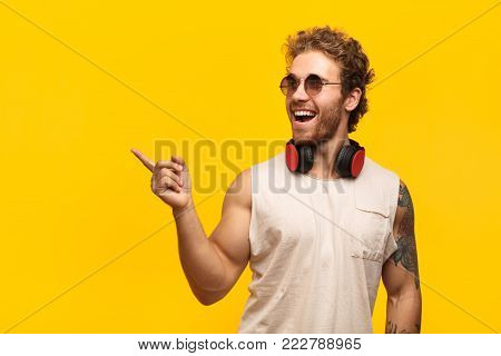 Young tattooed man in sunglasses and headphones posing cheerfully on orange background pointing away.