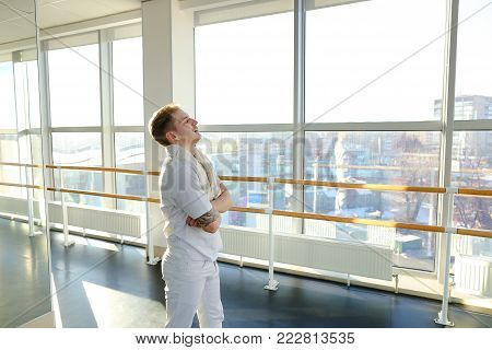 CEO of IT-company in sportswear discussing business on smartphone during training, young man receive important call in gym. Fair-haired smiling tattooed guy sitting on parquet floor speaking on mobile with partner making notes on planchette. Concept of in