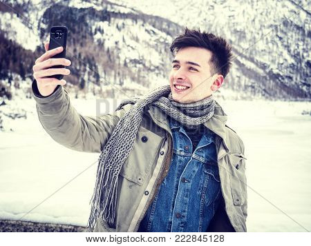 Handsome young man using smarpthone to take selfie photo, while posing among snowy woods in the mountain