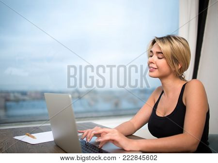 Charming woman website content creator working on modern laptop computer, sitting in office against window with copy space. Smiling female experiencer freelancer keyboarding on portable net-book