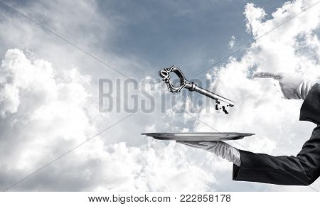 Cropped image of waitress's hand in white glove presenting big key on metal tray and pointing on it with cloudy skyscape on background.