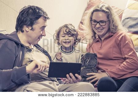 Happy family with baby girl sitting on a couch and using a digital tablet. Cheerful two-year-old child looks electronic device at home. Father and mother show something to their joyful little child on the tablet screen.