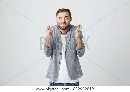 Supertitious handsome bearded European male dressed in gray jacket over white t-shirt crossing his fingers, wishful, hoping for better, looking upwards. Body language concept