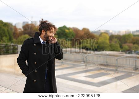 Half Nigerian guy near Eiffel Tower sends messages to girlfriend using modern smartphone in slow motion. Handsome jocund man wears black coat, ring with white stone and earring, has dark hair, beard and brown eyes. Concept of new apps for talking and text
