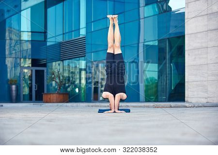 Young Man Practices Headstand Yoga Asana Sirsasana Pose Outdoors Against The Background Of A Modern 