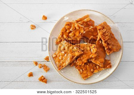Plate Of Traditional Peanut Brittle Candy Pieces. Top View On A White Wood Background.