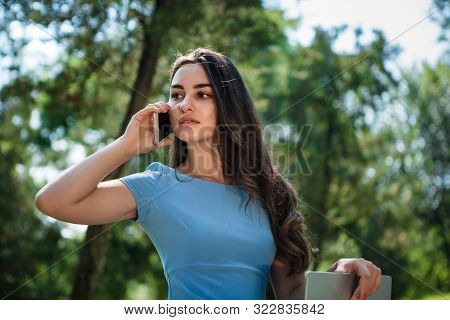 Young Brunette Srudent In A Blue Dress Sitting With A Laptop In A Park And Talking On The Phone. Con