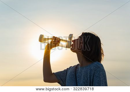 Young Woman Drinks Water From A Bottle On A Sunset Sky Background. Portrait Of A Female With A Reusa