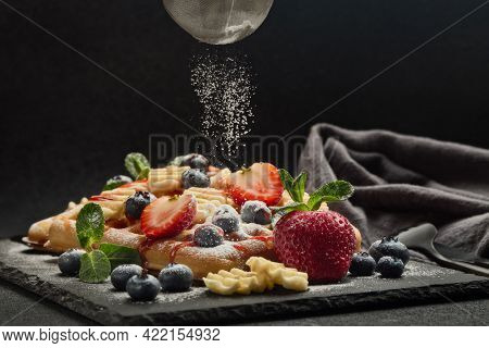 Delicious waffles with berries, mint, powdered sugar, sweet sauce on slate serving board on dark background.  Dessert. Serving food. Process of sprinkling icing sugar over waffles