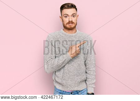 Young redhead man wearing casual winter sweater pointing aside worried and nervous with forefinger, concerned and surprised expression 