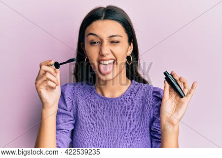 Young hispanic woman holding eyelashes curler sticking tongue out happy with funny expression. 
