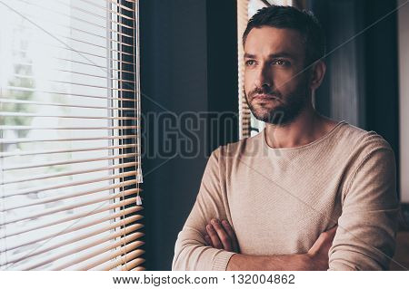 Handsome and thoughtful. Handsome young man looking trough window while standing at his working place