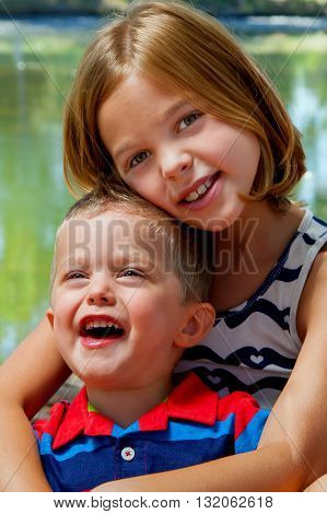 A little brother sits in his older sister's lap as he laughs at someone in the distance. His siter smiles while looking at the camera.
