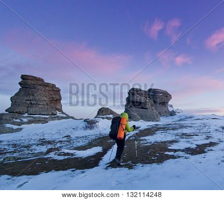 man with backpack and poles going in mountains and rocks snow covered at purple sunset