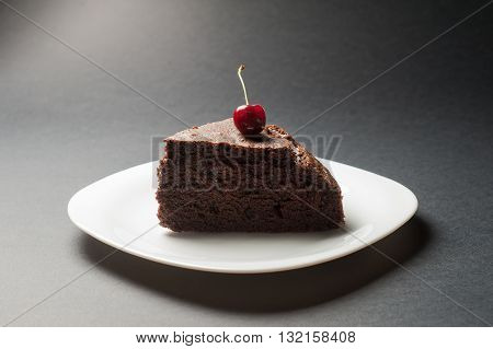 Slice of delicious chocolate cake with cherry on top on a white plate on a dark background.