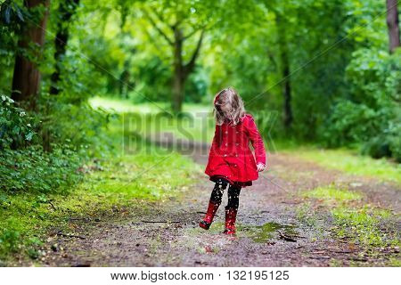 Little girl playing in rainy summer park. Child with red ladybug umbrella waterproof coat and boots jumping in puddle and mud in the rain. Kid walking in autumn shower. Outdoor fun by any weather.