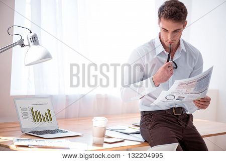 Get the information. Pleasant concentrated brutal man leaning on the table and reading newspaper while standing in the office