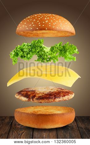 Burger ingredients. Burger layers isolated. Separated burger ingredients on wood. Hamburger explosion. Cheeseburger flying layers on brown background. Levitation of burger and cheese, meat, lettuce.