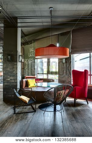 Room in a mexican restaurant in a loft style. There is a glass rounded table with black chairs, a crimson armchair and a brown sofa. Over them there is a red lampshade. Multi-colored pillows lies on the sofa and the chairs. 