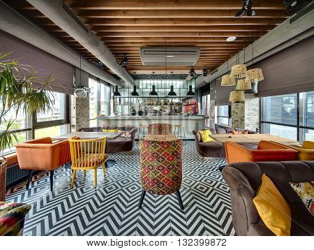 Room in a loft style in a mexican restaurant with open kitchen on the background. There are wooden tables with brown sofas and multi-colored chairs. On the sofas there are color pillows. On the sides there are brick walls with windows with curtains. 