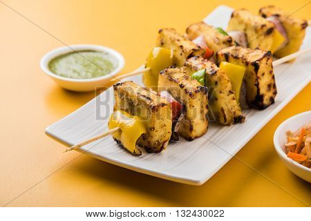 Paneer Tikka Kabab - Tandoori Indian cheese skewers, malai paneer tikka / malai paneer kabab, chilli paneer served in white plate with barbecue stick and colourful capsicum and onion, with green sauce