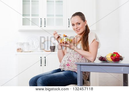 Pregnant woman eats fruit salad on the house kitchen.