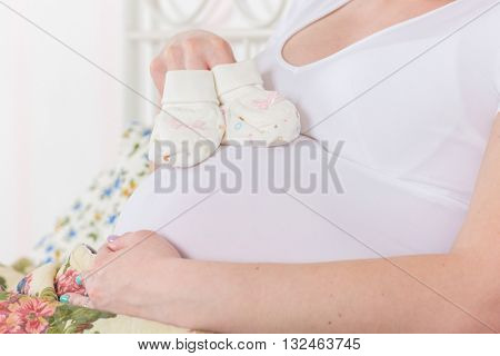 Pregnant woman with children's bootees  sits on a bed in the room.
