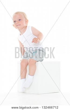 Unhappy yearling baby boy sits on cube on a white background.
