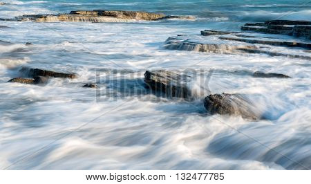 Sea waves crashing on plates of sea rocks creating small waterfalls and smooth streams of water.