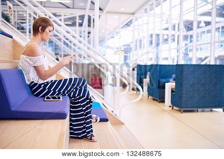 Young caucasian female creative with short brwon hair sitting on a colourful cushion in a co working space while texting about business on her smartphone, with her tablet by her side.