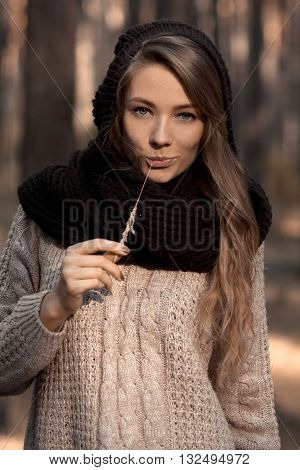 Close-up of fashionable model, glamorous girl in scarf and hat with straw in teeth, fingers, hands, mouth. Perfect sunny face with blue eyes. Sunny portrait of smiling girl. Creative idea. Autumn forest.