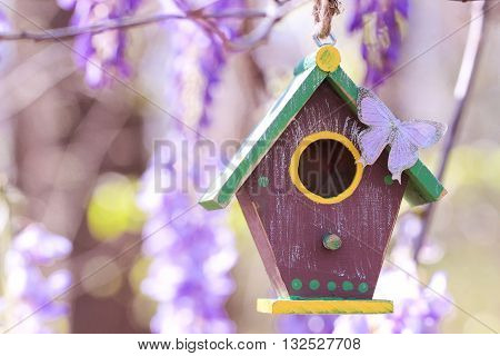 Brown and green birdhouse with purple butterfly hanging from tree branch with purple spring flowers
