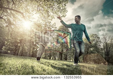 Runnings little boy and father flies with them kite in the park under sunligt.