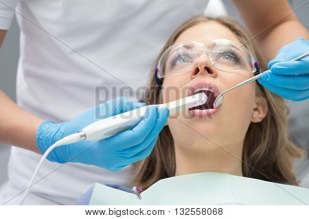 Pretty girl with opened mouth in patient bib and protective eyewear. Next to her there is a dentist in a white uniform with blue latex gloves. He diagnoses her teeth with a LED intraoral camera and a stem mouth mirror. Horizontal.
