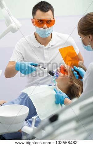 Young girl in blue shirt and patient bib on the patient chair in the dental cabinet. Next to her there is a male dentist and a female assistant. They both wear white uniform with blue latex gloves and blue masks. Patient and dentist wear UV protective eye