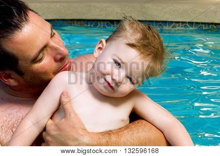 A father holds his toddler son in a swimming pool on a summer day. The boy's blue eyes match the color of the water. The boy has ptosis.