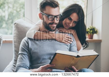 Reading their favorite book. Beautiful young woman bonding to her husband while he is reading a book on the couch