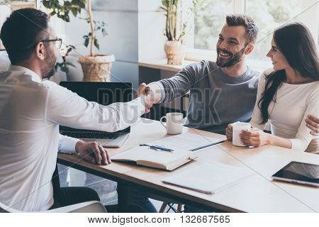 Sealing a deal. Cheerful young man bonding to his wife while shaking hand to man sitting in front of him at the desk