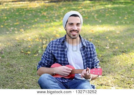 Portrait of young handsome man playing the ukelele in a park. Outdoors.