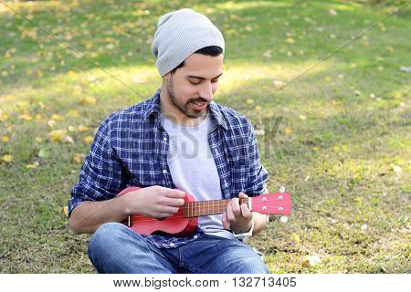 Portrait of young handsome man playing the ukelele in a park. Outdoors.