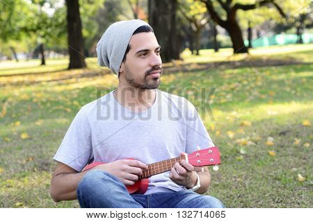 Portrait of young handsome man playing the ukelele in a park. Outdoors.