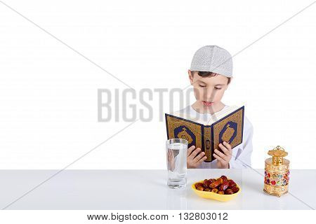 Young Muslim boy reading Quraan in Ramadan - sitting at a table with water dates Quran and Ramadan lantern