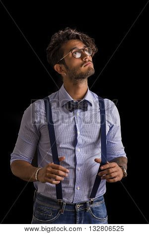 Portrait of brunette young man in glasses, bow-tie, suspenders and shirt looking at camera. Studio shot isolated on black background