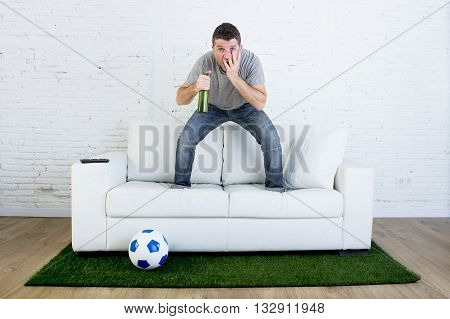 crazy football fan cheering watching television soccer match suffering stress nervous and excited jumping on sofa couch with ball on grass carpet emulating stadium pitch
