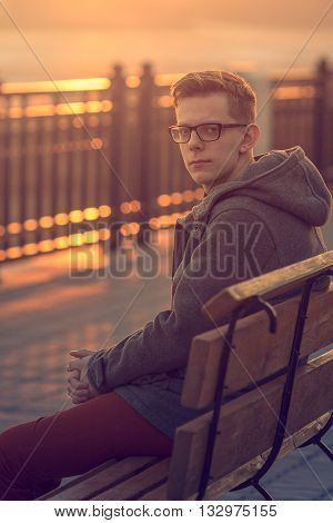 Photo of Lonely Man Sitting on a Bench during Sunset. Portrait of Young Man Sitting on Wooden Bench. Sunset Male Outdoor Portrait