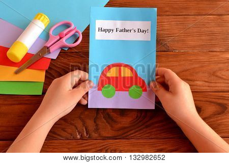 Child holds a greeting card in his hands. Kids crafts for father's day. Card with message Happy father's day. Holiday gift for dad. Sheets of colored paper, scissors, glue stick. Wooden background