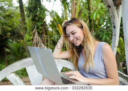 Happy Girl Using Laptop And Sitting In Exotic Garedn With Palms In Background.