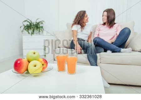 Pile Of Fresh Yummy Apples And Two Glasses Of Juice On The Table On The Forefront. Two Amazing Girls