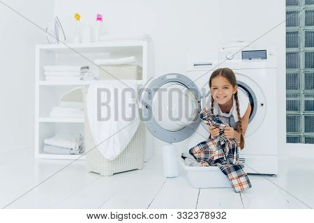 Positive Kid With Pigtails Sticks Out Head From Washing Machine, Has Fun And Holds Shirt, Prepares F
