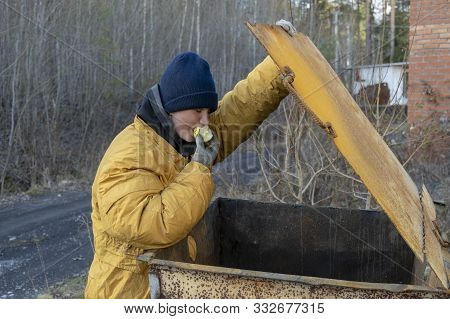 A Homeless Woman In A Yellow Old Ragged Jacket And A Blue Hat Opens A Tank And Rummages Through The